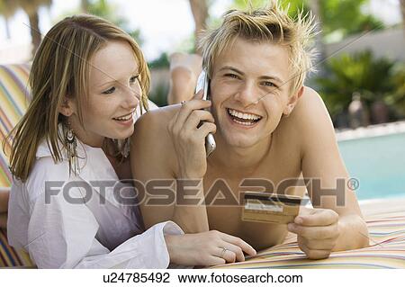 Stock Image - Young couple on deck chair by pool, man making credit card purchase on mobile phone, portrait. Fotosearch