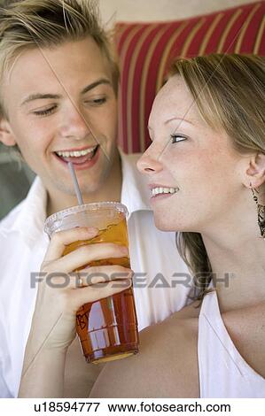 Stock Photo - Young couple on sofa, woman giving man iced drink, close up. Fotosearch