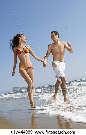 Stock Photo - Young couple running through surf on beach, front view. Fotosearch