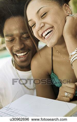 Stock Photograph - Young couple sitting at back yard table using laptop and laughing, close up. Fotosearch