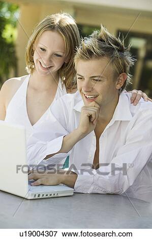 Stock Photo - Young couple sitting at backyard table using laptop. Fotosearch
