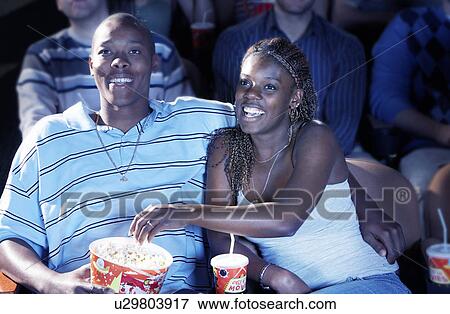 Young Couple sitting in theatre, arms around, Watching Movie, eating popcorn View Large Photo Image Stock Photo - Young Couple sitting in theatre, arms around, Watching Movie, eating popcorn. Fotosearch