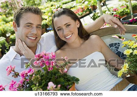 Stock Image - Young couple sitting on bench in garden centre, (portrait). Fotosearch