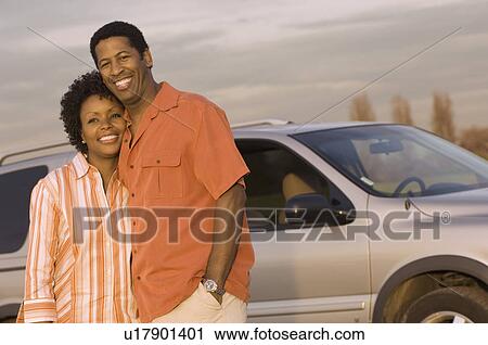 Stock Image - Young couple standing together in front of car, half-length. Fotosearch