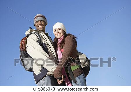 Stock Image - Young couple wearing backpacks, standing outdoors holding hands.. Fotosearch