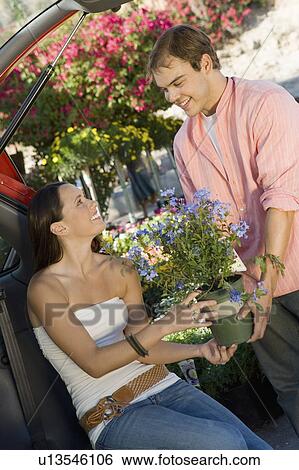 Stock Photograph - Young couple with potted flower in garden centre, woman sitting in open car trunk. Fotosearch