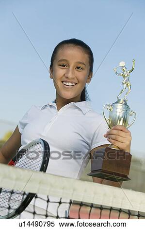 Stock Photography - Young girl at net on tennis court holding trophy, portrait, low angle view. Fotosearch