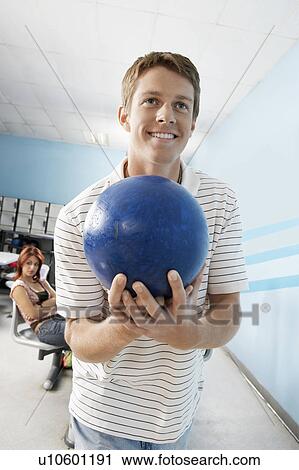 Stock Image - Young man at bowling alley holding ball, portrait. Fotosearch