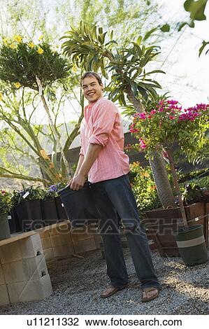 Stock Image - Young man holding small tree in pot in garden centre, smiling. Fotosearch
