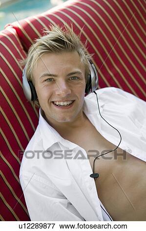Young man on deckchair by pool listening to music on headphones, portrait View Large Photo Image Stock Photo - Young man on deckchair by pool listening to music on headphones, portrait. Fotosearch