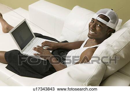 Stock Image - Young man on sofa using laptop, high angle view, portrait. Fotosearch