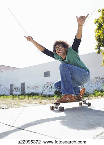Stock Image - Young man with arms raised on skateboard, outdoors, portrait. Fotosearch