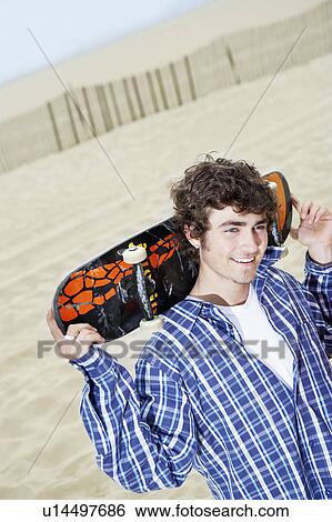 Stock Photograph - Young man with skateboard on shoulders on beach. Fotosearch