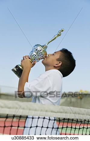 Stock Image - Young tennis player by net on court kissing trophy, side view. Fotosearch