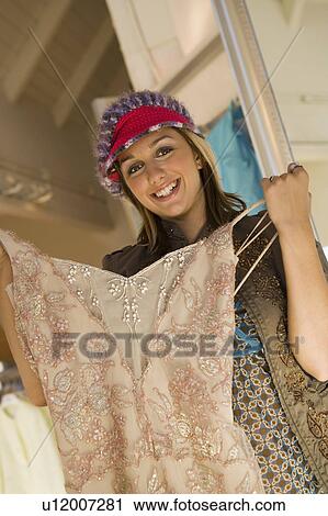 Stock Image - Young woman holding dress in shop, (portrait). Fotosearch