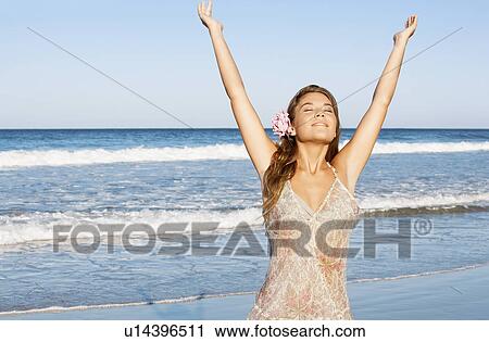 Stock Image - Young woman in summer dress by sea, arms raised. Fotosearch