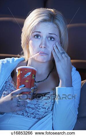 Stock Photograph - Young woman sitting in theatre, drinking soft drink, hand on cheek, watching sad movie. Fotosearch