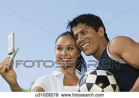 Stock Photo - Young woman using mobile phone, photographing self with young man.. Fotosearch