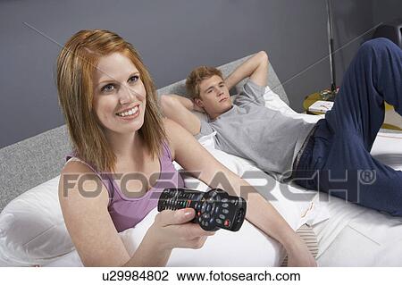 Stock Image - Young woman using remote control lying in bed next to boyfriend. Fotosearch