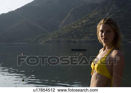 Stock Image - Blonde woman in bikini looks back at camera in morning sunlight of lake. Fotosearch