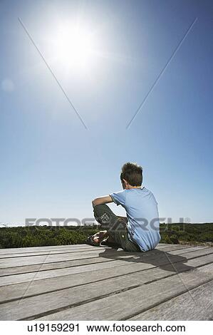 Stock Image - Boy sitting on boardwalk, back view. Fotosearch