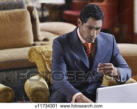 Business man sitting in lobby working on laptop, holding eyeglasses View Large Photo Image Stock Image - Business man sitting in lobby working on laptop, holding eyeglasses. Fotosearch