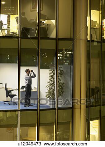 Stock Image - Business man using binoculars in office, view from building exterior. Fotosearch