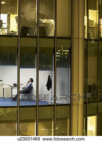Stock Photo - Business man working at desk in office, view from building exterior. Fotosearch