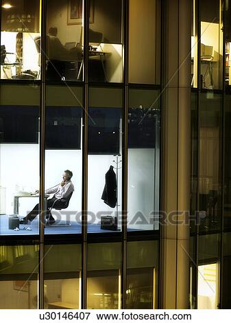 Stock Photo - Business man working at desk in office, view from building exterior. Fotosearch