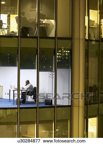 Stock Photo - Business man working at desk in office, view from building exterior. Fotosearch