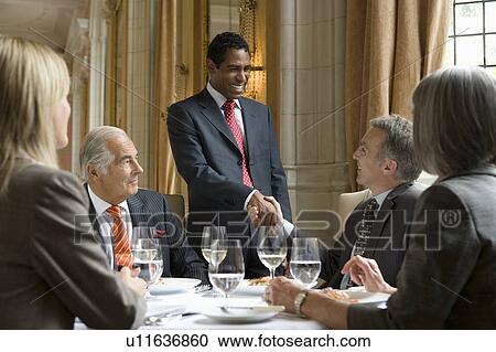 Stock Image - Business people at meeting at restaurant, one man standing. Fotosearch