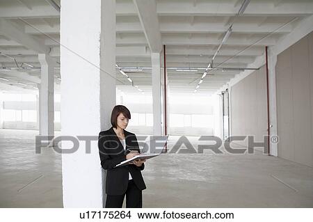 Picture - Business woman leans against pillar with file in empty warehouse. Fotosearch