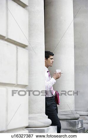 Stock Photo - Businessman leaning on pillar outside building holding coffee. Fotosearch
