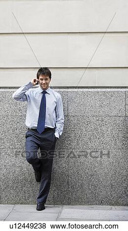 Stock Photo - Businessman on cell phone leaning on wall of building. Fotosearch