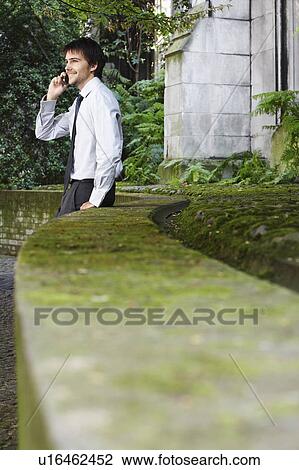 Businessman on cell phone leaning against wall in park View Large Photo Image Stock Image - Businessman on cell phone leaning against wall in park. Fotosearch