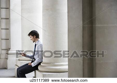 Stock Photo - Businessman outside building sitting on pillar sending text message. Fotosearch