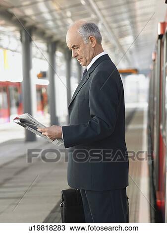 Stock Image - Businessman Reading Newspaper at empty Train Station, side view. Fotosearch