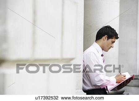 Stock Image - Businessman sitting against pillar outside building writing in notebook. Fotosearch