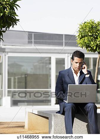 Businessman sitting alone in plaza using laptop and cell phone View Large Photo Image Stock Photography - Businessman sitting alone in plaza using laptop and cell phone. Fotosearch