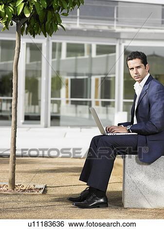 Businessman sitting in plaza using laptop View Large Photo Image Stock Photograph - Businessman sitting in plaza using laptop. Fotosearch