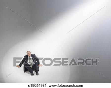Stock Image - Businessman sitting, looking up at source of spotlight. Fotosearch
