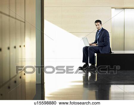 Stock Image - Businessman sitting on bench by elevator using laptop. Fotosearch