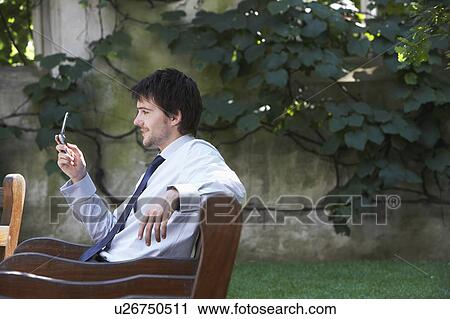 Stock Image - Businessman sitting on park bench sending text message. Fotosearch