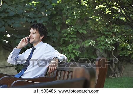Businessman sitting on park bench talking on cell phone View Large Photo Image Stock Photography - Businessman sitting on park bench talking on cell phone. Fotosearch