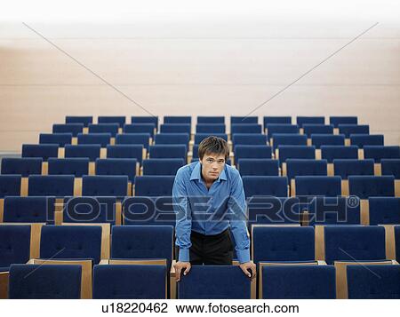 Stock Image - Businessman standing alone leaning on chair in Auditorium, portrait. Fotosearch