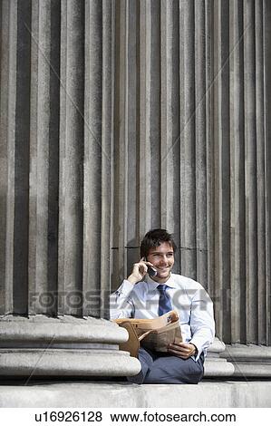 Businessman with newspaper sitting on pillar outside building talking on cell phone View Large Photo Image Stock Photo - Businessman with newspaper sitting on pillar outside building talking on cell phone. Fotosearch