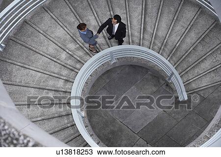 Stock Image - China, Hong, Kong, two business people shaking hands standing on spiral staircase, view from above. Fotosearch