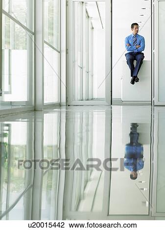 Stock Image - Confident Businessman sitting on desk in office, viewed through doorway. Fotosearch