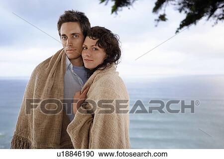 Stock Image - Couple covered by blanket standing by sea, portrait. Fotosearch