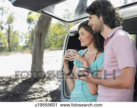 Stock Image - Couple drinking from thermos cups leaning in open tailgate of van, half length. Fotosearch